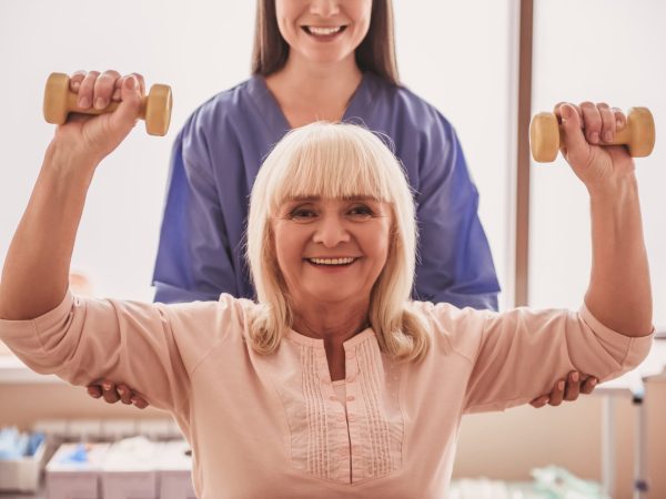 Beautiful old lady is doing exercises with dumbbells, looking at camera and smiling, in hospital ward. Attractive nurse is helping her