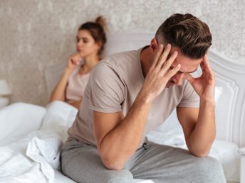Sad man sitting on bed with his girlfriend on background at home