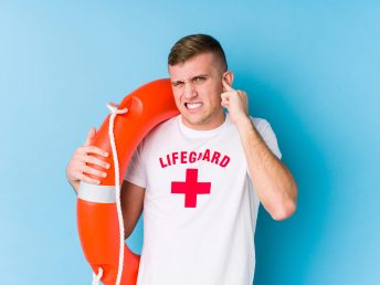 Young lifeguard man holding a rescue float covering ears with hands.